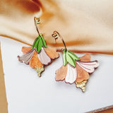 A pair of handcrafted Hibiscus Earrings with gold hoops, featuring pink, peach, and green petal designs, displayed on a white textured surface with beige satin fabric in the background.