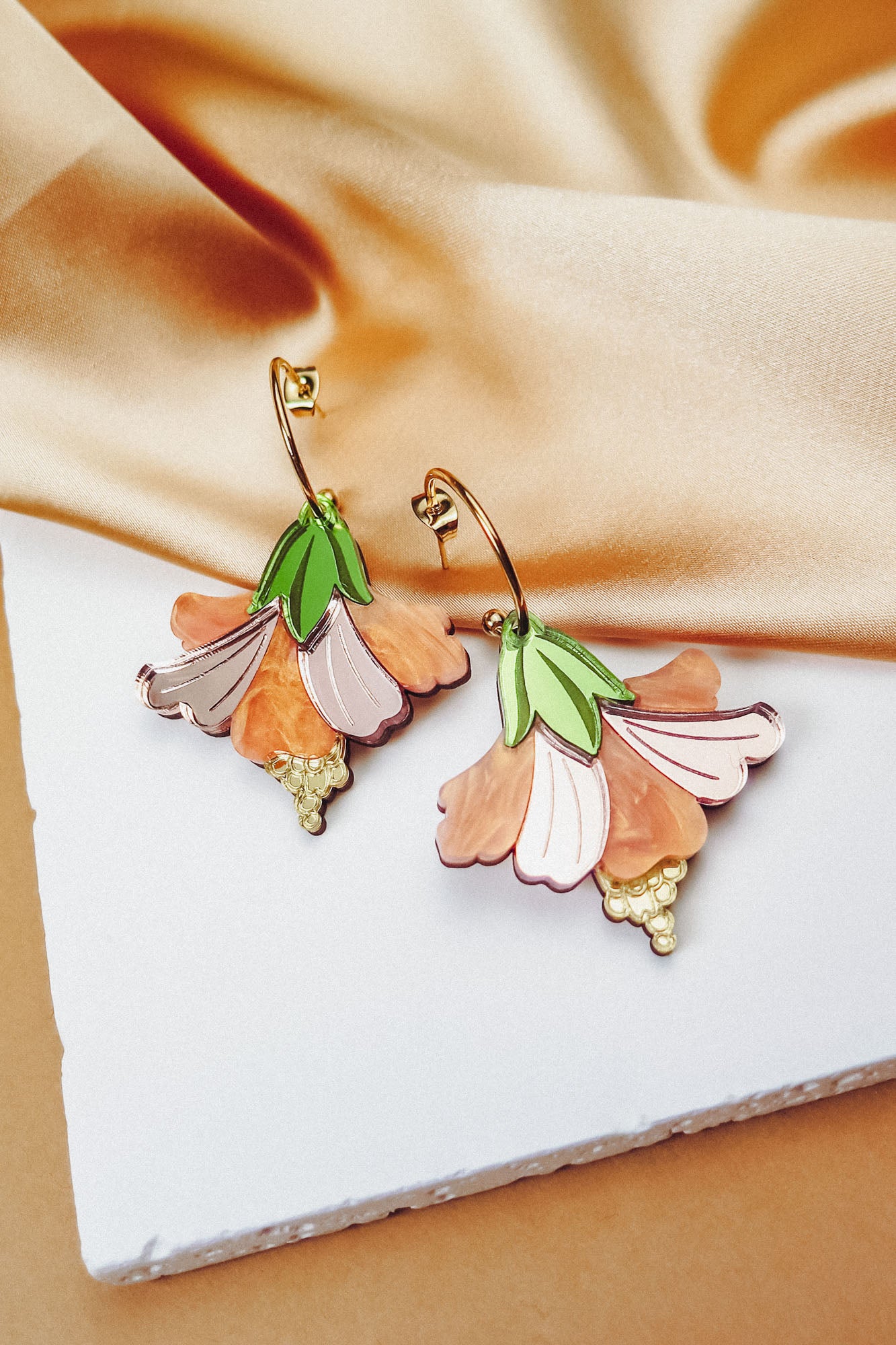 A pair of handcrafted Hibiscus Earrings with gold hoops, featuring pink, peach, and green petal designs, displayed on a white textured surface with beige satin fabric in the background.