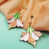 A pair of colorful, handcrafted Hibiscus Earrings with gold hooks rest on peach-colored fabric and an aqua background. Featuring pink, peach, and green hues with gold bead accents, they make a perfect tropical statement accessory.