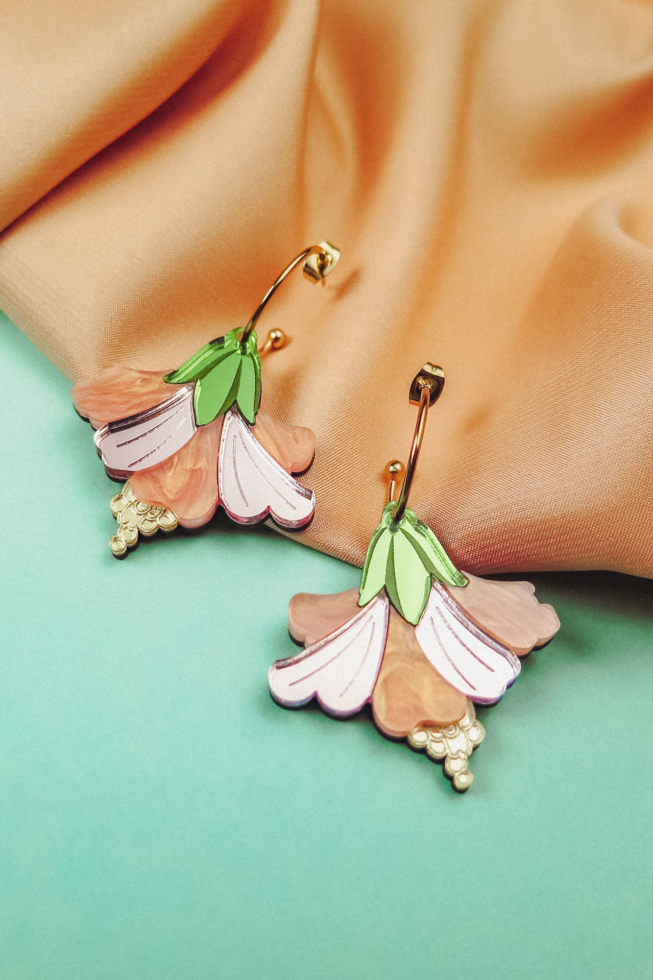 A pair of colorful, handcrafted Hibiscus Earrings with gold hooks rest on peach-colored fabric and an aqua background. Featuring pink, peach, and green hues with gold bead accents, they make a perfect tropical statement accessory.