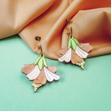 A pair of handcrafted Hibiscus Earrings, shaped like pastel-colored flowers with gold accents, are displayed on a pastel blue surface with peach-colored fabric in the background.