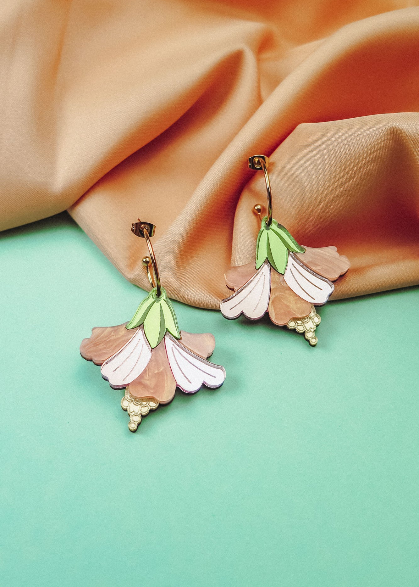 A pair of handcrafted Hibiscus Earrings, shaped like pastel-colored flowers with gold accents, are displayed on a pastel blue surface with peach-colored fabric in the background.
