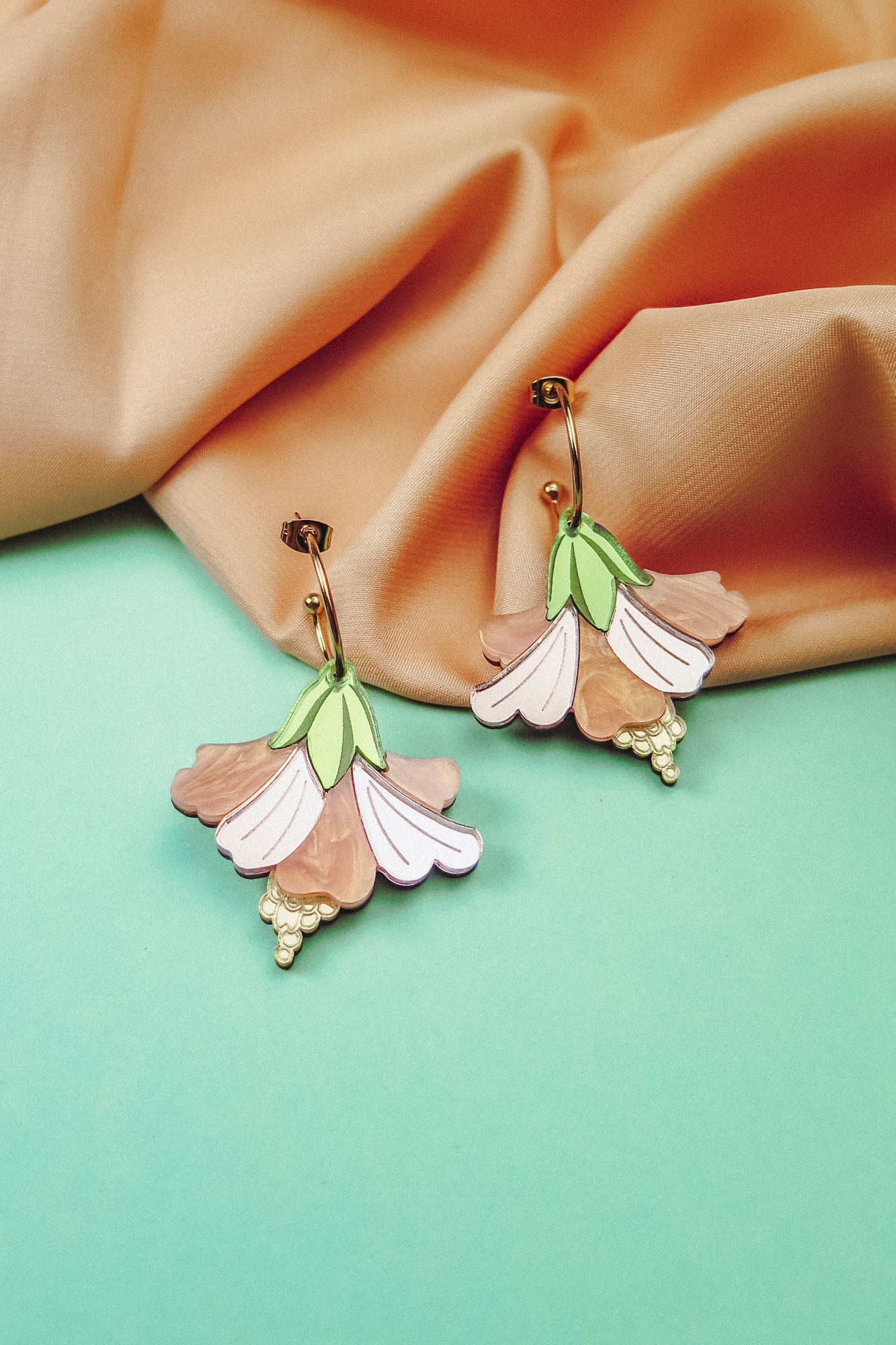 A pair of handcrafted Hibiscus Earrings, shaped like pastel-colored flowers with gold accents, are displayed on a pastel blue surface with peach-colored fabric in the background.