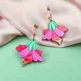 Handcrafted Hibiscus Earrings in a tropical statement style, featuring colorful pink and red petals, green leaves, and gold accents, are displayed on soft pink fabric and a mint green background.
