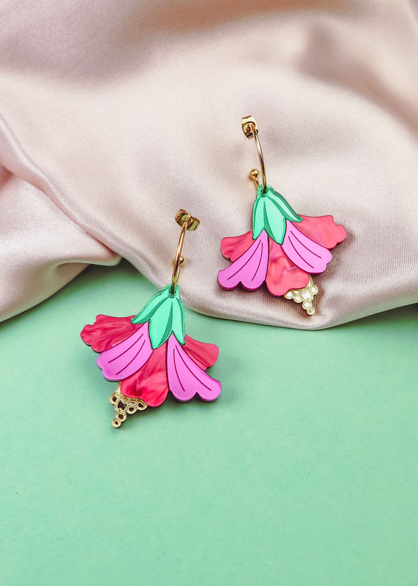 Handcrafted Hibiscus Earrings in a tropical statement style, featuring colorful pink and red petals, green leaves, and gold accents, are displayed on soft pink fabric and a mint green background.