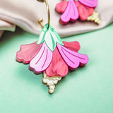 A close-up of handcrafted Hibiscus Earrings, featuring a tropical statement flower with pink and magenta petals, green leaves, and gold-tone details, resting on a mint green surface near soft beige fabric.