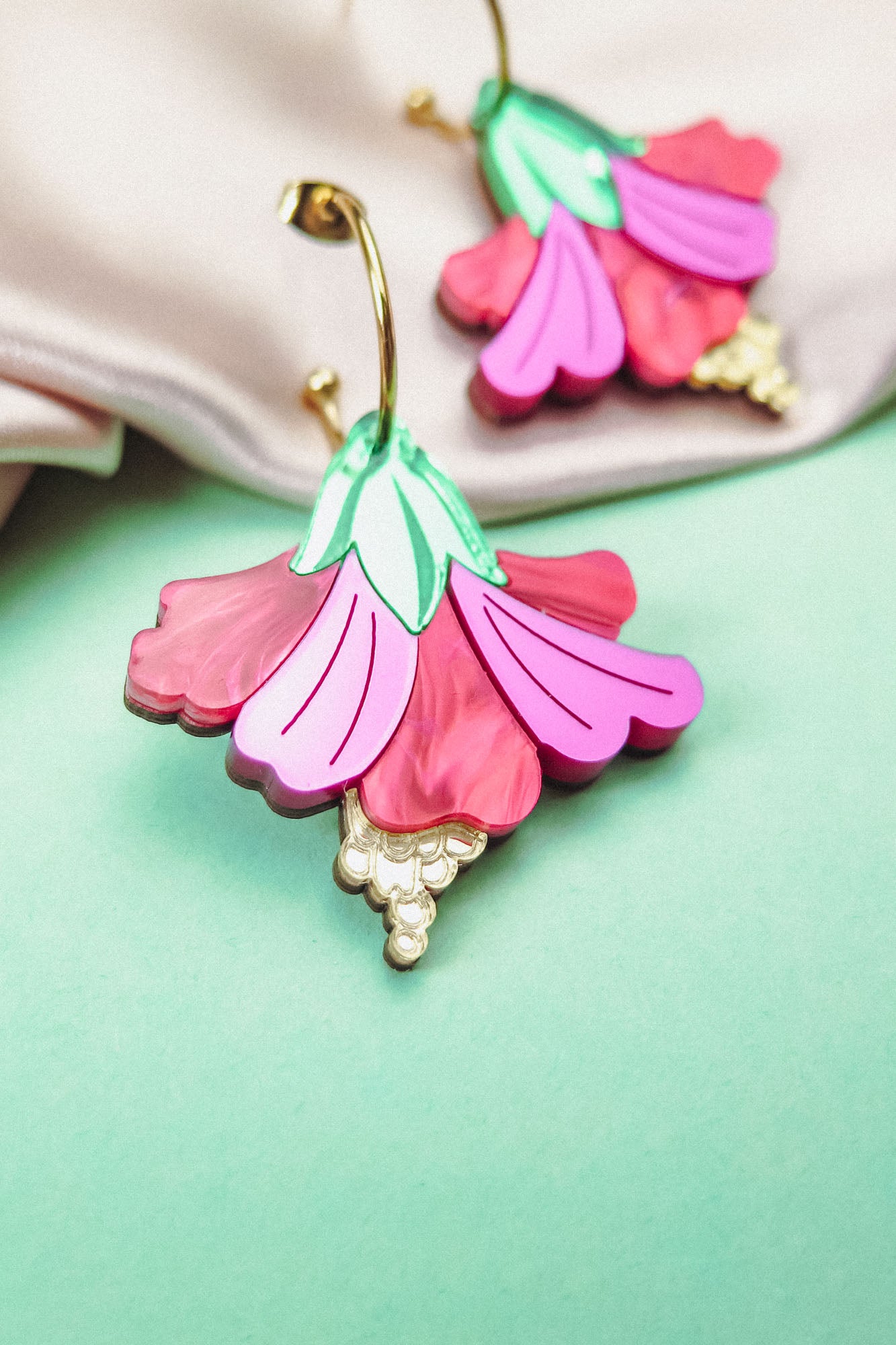 A close-up of handcrafted Hibiscus Earrings, featuring a tropical statement flower with pink and magenta petals, green leaves, and gold-tone details, resting on a mint green surface near soft beige fabric.