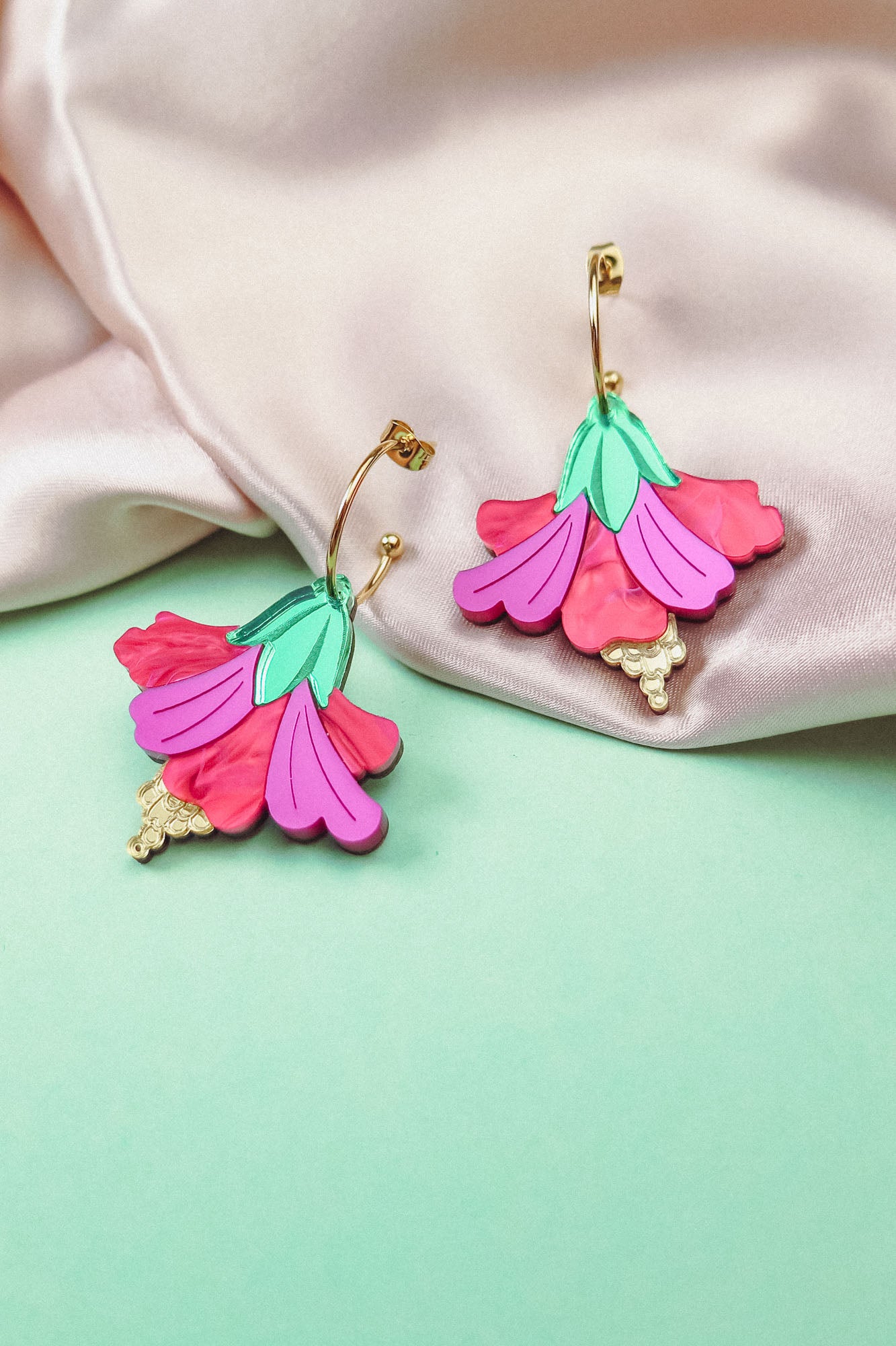 Two colorful Hibiscus Earrings with gold hoops and pink, purple, and green petal designs rest on light pink satin fabric and a mint green surface, making a handcrafted tropical statement.