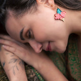A woman with long brown hair lies on her side outdoors, displaying vibrant Hibiscus Earrings and a botanical tattoo on her arm. She is wearing a green patterned garment and appears calm and relaxed, making a tropical statement.