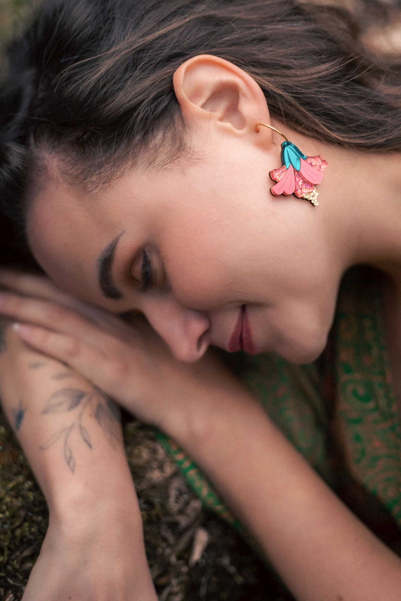 A woman with long brown hair lies on her side outdoors, displaying vibrant Hibiscus Earrings and a botanical tattoo on her arm. She is wearing a green patterned garment and appears calm and relaxed, making a tropical statement.