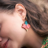 A woman with wavy brown hair wears vibrant Hibiscus Earrings—gold hoops adorned with pink, teal, and gold floral charms. She is smiling softly, her tropical statement green patterned clothing visible.