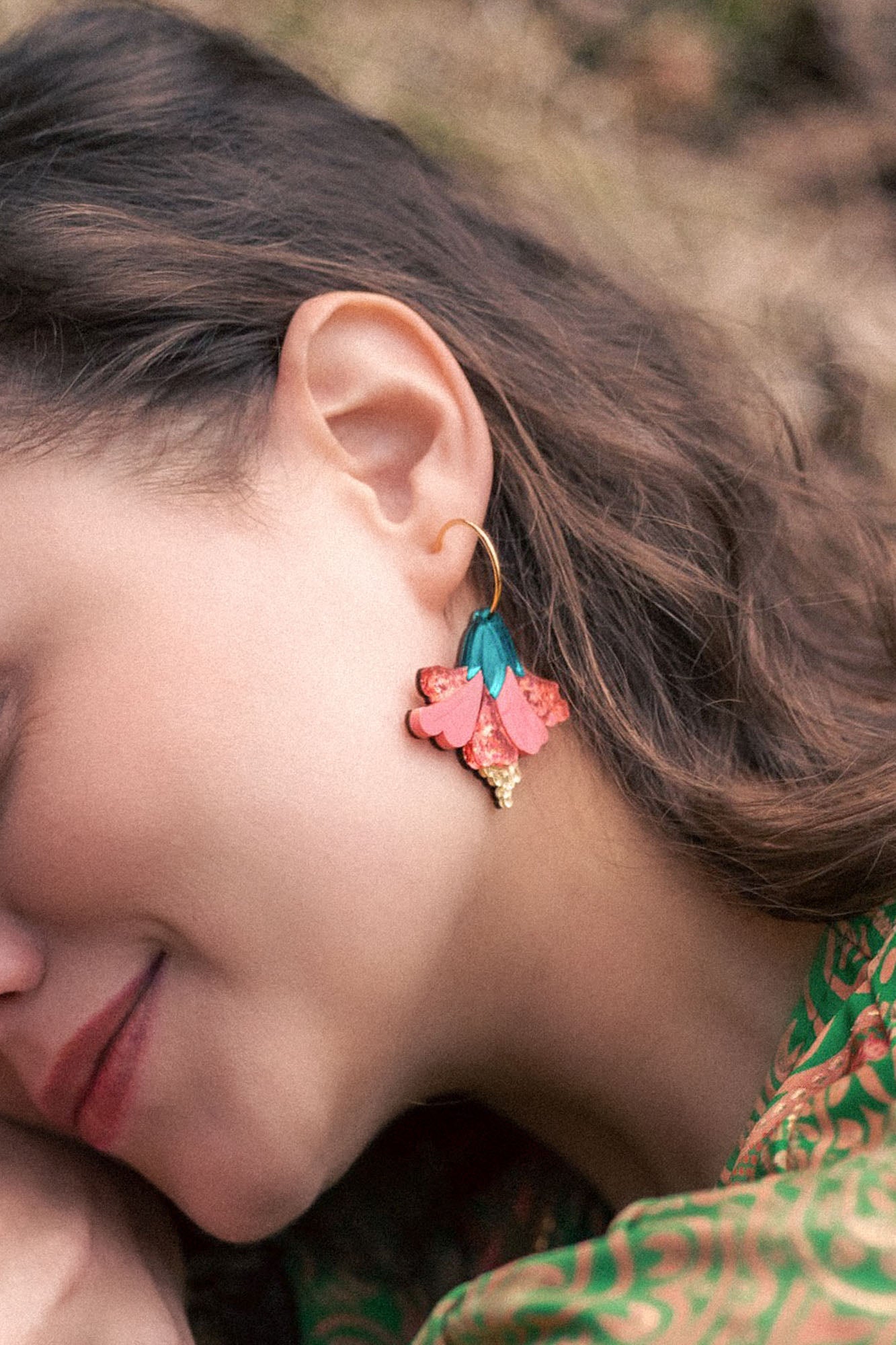 A woman with wavy brown hair wears vibrant Hibiscus Earrings—gold hoops adorned with pink, teal, and gold floral charms. She is smiling softly, her tropical statement green patterned clothing visible.