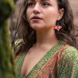 A woman with wavy brown hair wears vibrant, butterfly-shaped earrings and a green patterned dress, standing outdoors near moss-covered branches, looking thoughtfully into the distance—a true tropical statement.