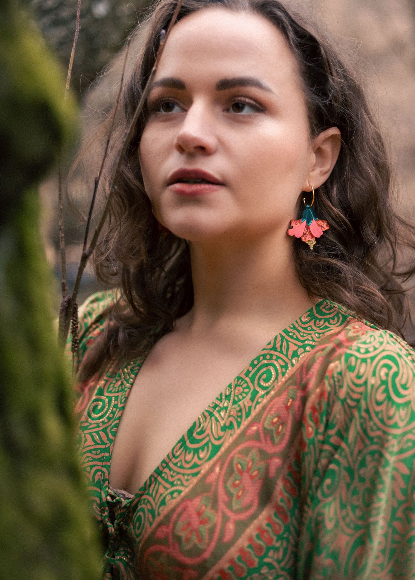 A woman with wavy brown hair wears vibrant, butterfly-shaped earrings and a green patterned dress, standing outdoors near moss-covered branches, looking thoughtfully into the distance—a true tropical statement.