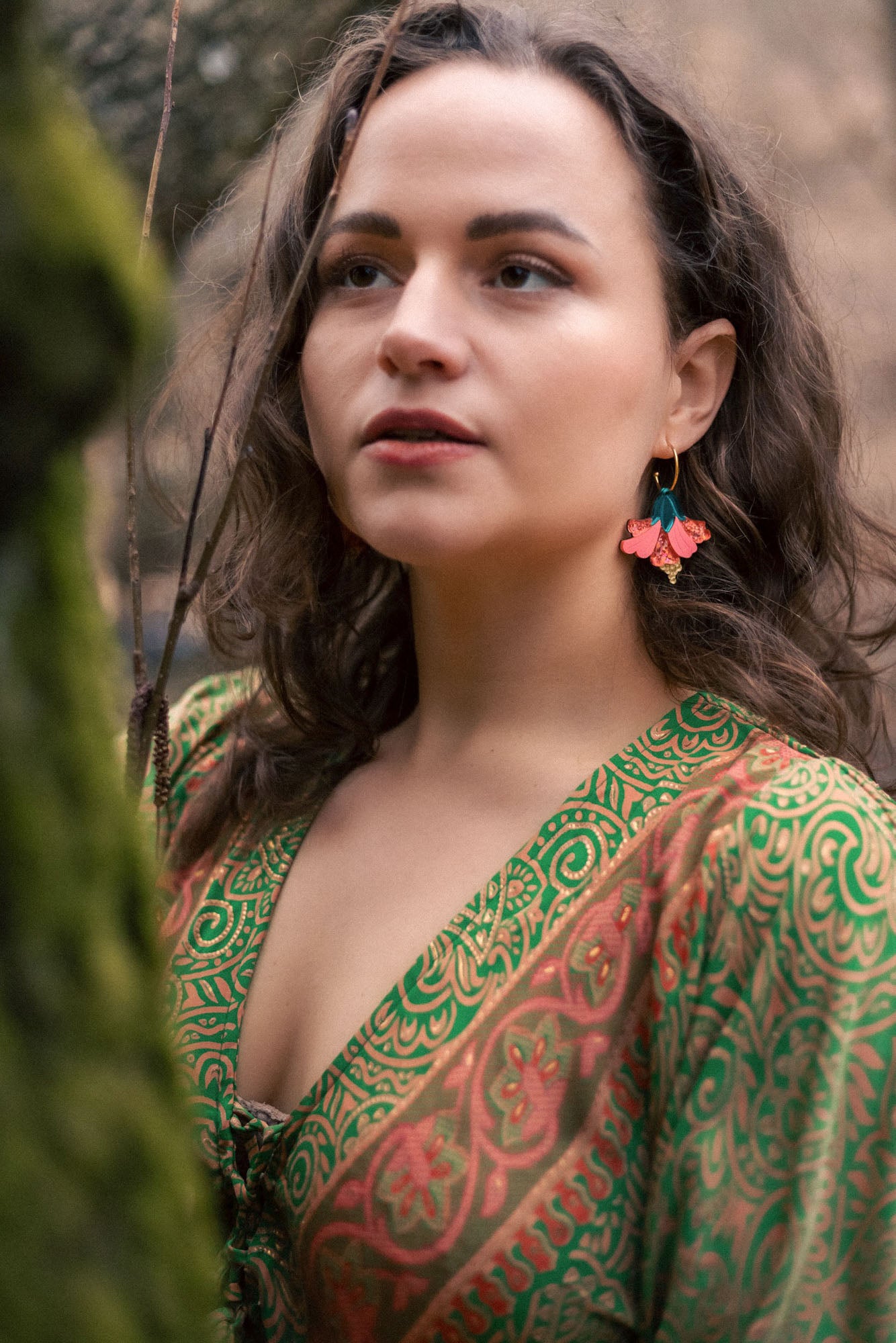 A woman with wavy brown hair wears vibrant, butterfly-shaped earrings and a green patterned dress, standing outdoors near moss-covered branches, looking thoughtfully into the distance—a true tropical statement.