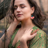 A woman with wavy brown hair and closed eyes stands outdoors, wearing a green patterned dress and vibrant Hibiscus Earrings, touching her face and neck gently, with a tattoo visible on her forearm.