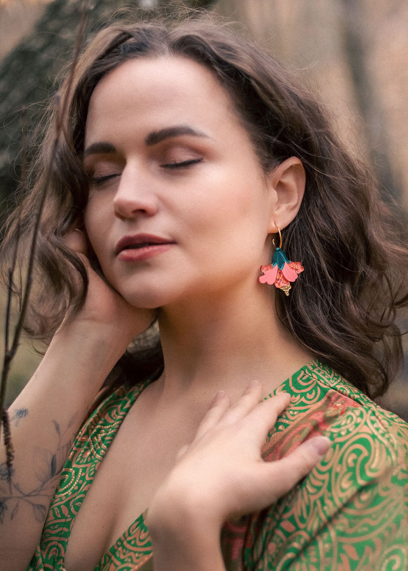 A woman with wavy brown hair and closed eyes stands outdoors, wearing a green patterned dress and vibrant Hibiscus Earrings, touching her face and neck gently, with a tattoo visible on her forearm.