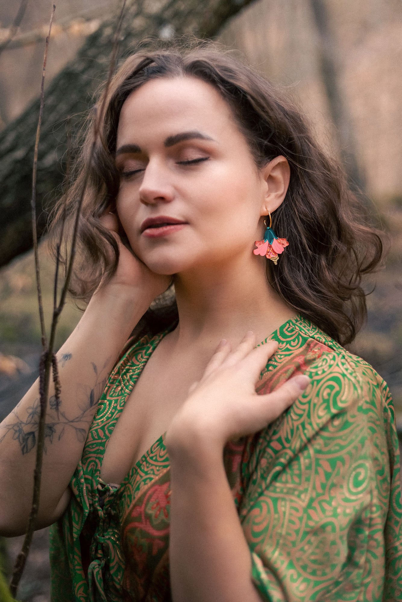 A woman with wavy brown hair and closed eyes stands outdoors, wearing a green patterned dress and vibrant Hibiscus Earrings, touching her face and neck gently, with a tattoo visible on her forearm.