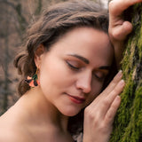 A woman with wavy brown hair and handcrafted Hibiscus Earrings gently leans her head and hand against a mossy tree, eyes closed, wearing an off-shoulder blouse—radiating a tropical statement in a serene forest setting.