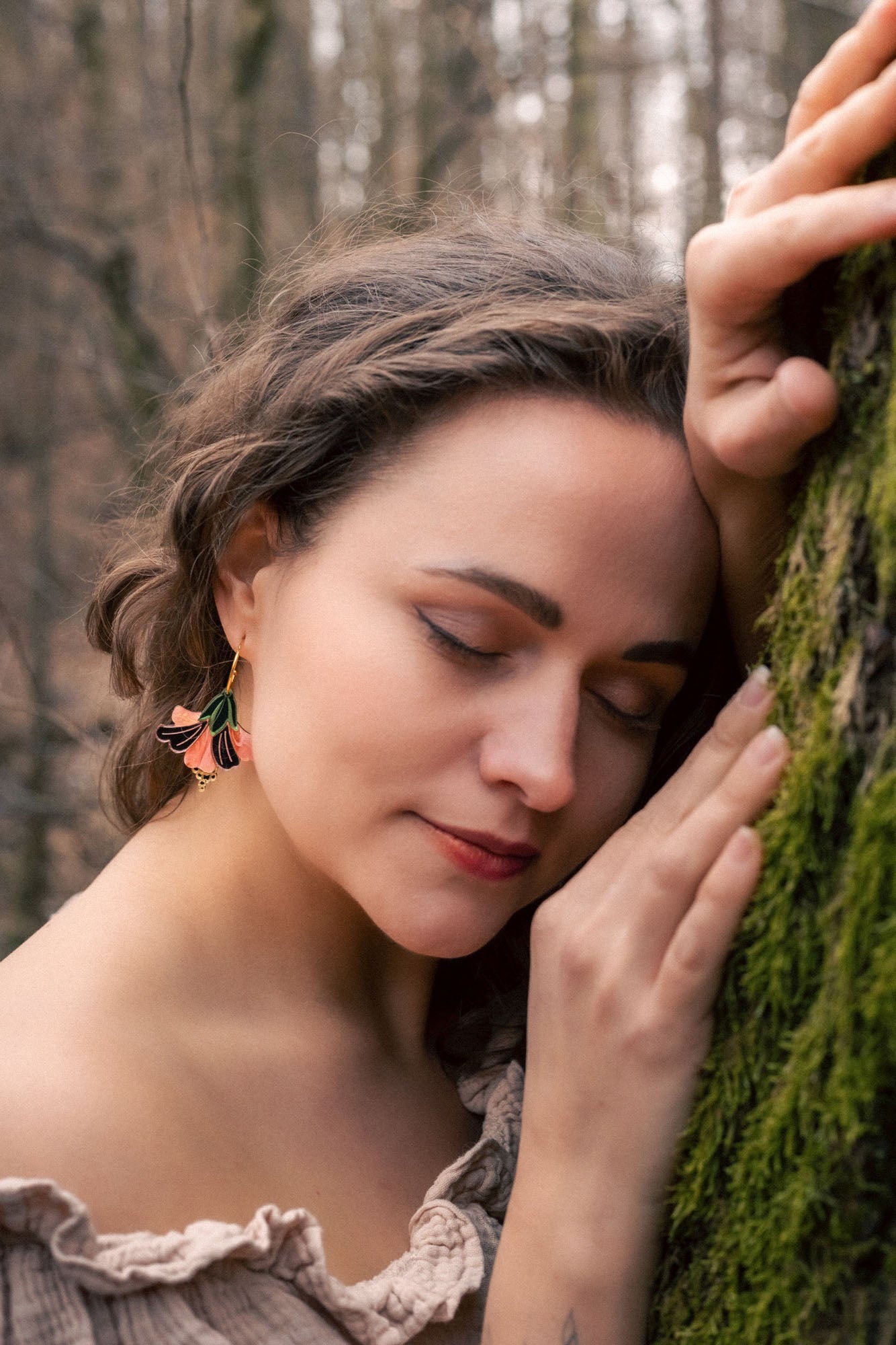 A woman with wavy brown hair and handcrafted Hibiscus Earrings gently leans her head and hand against a mossy tree, eyes closed, wearing an off-shoulder blouse—radiating a tropical statement in a serene forest setting.
