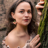 A woman with wavy brown hair and floral handcrafted Hibiscus Earrings stands in a forest, wearing an off-shoulder beige top. She gently holds a mossy tree trunk and looks at the camera with a soft, calm expression.