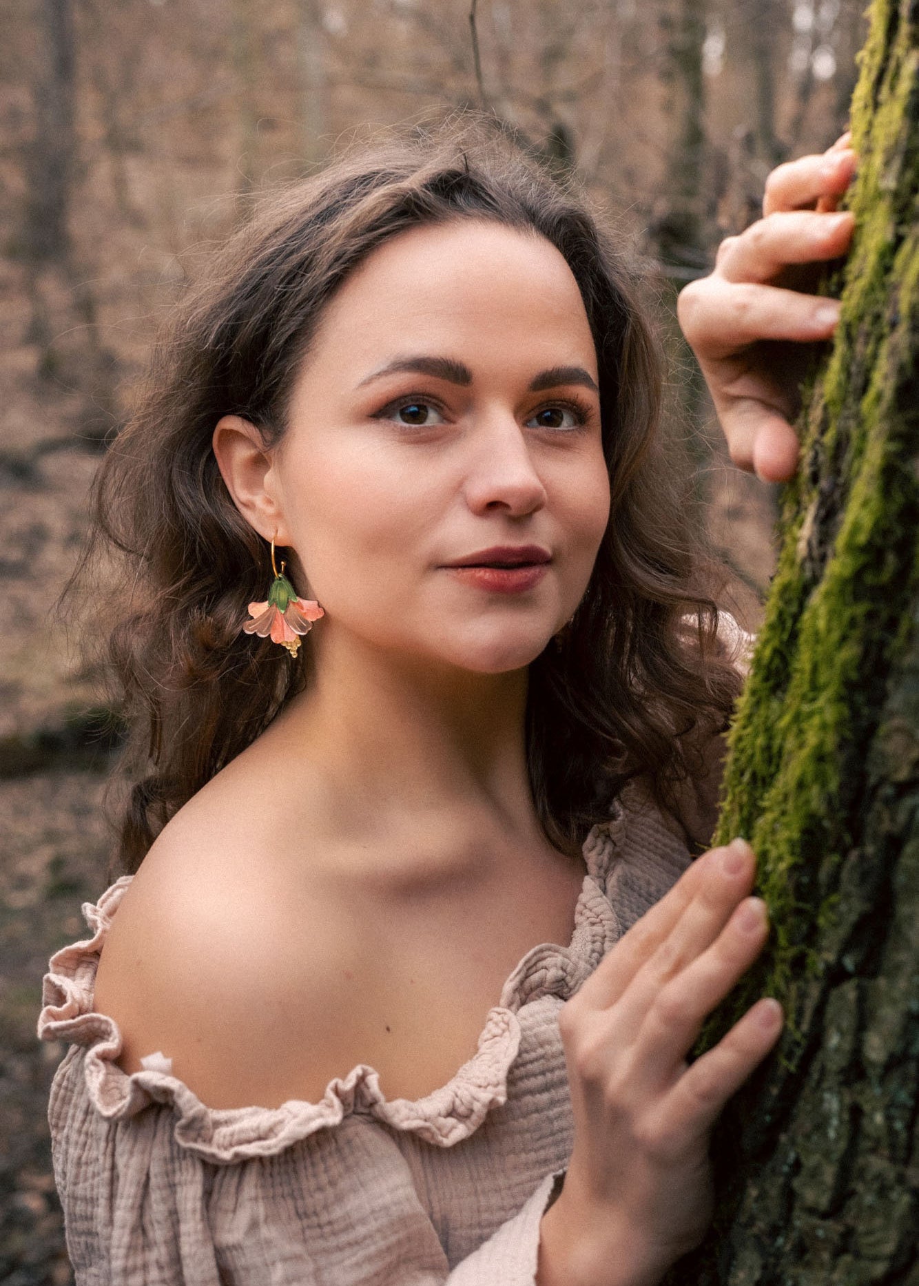 A woman with wavy brown hair and floral handcrafted Hibiscus Earrings stands in a forest, wearing an off-shoulder beige top. She gently holds a mossy tree trunk and looks at the camera with a soft, calm expression.