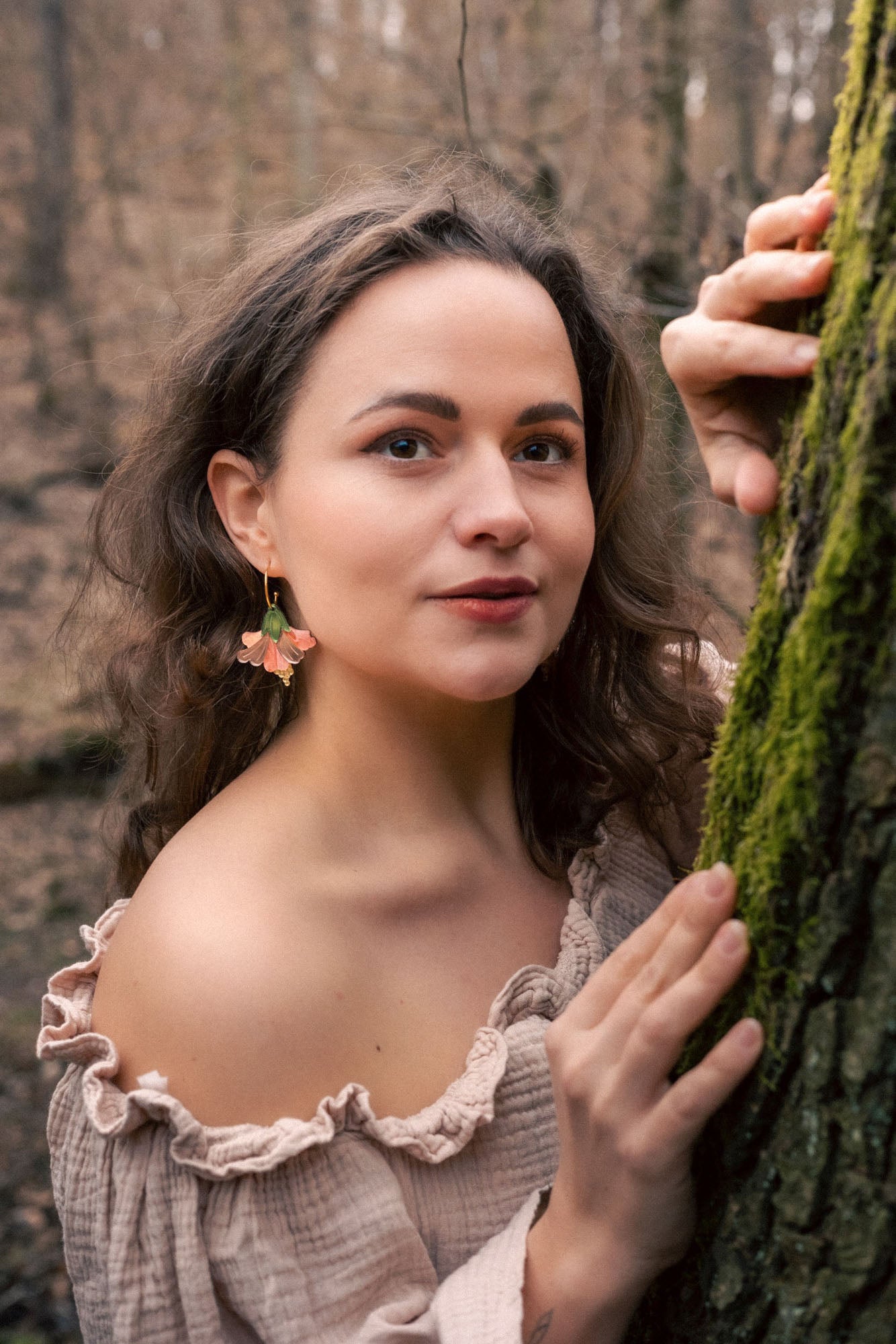 A woman with wavy brown hair and floral handcrafted Hibiscus Earrings stands in a forest, wearing an off-shoulder beige top. She gently holds a mossy tree trunk and looks at the camera with a soft, calm expression.