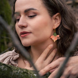 A woman with wavy brown hair and handcrafted Hibiscus Earrings gently touches her neck while standing in a forest. Her eyes are closed, branches cross before her, and she wears a beige top with ruffled details.