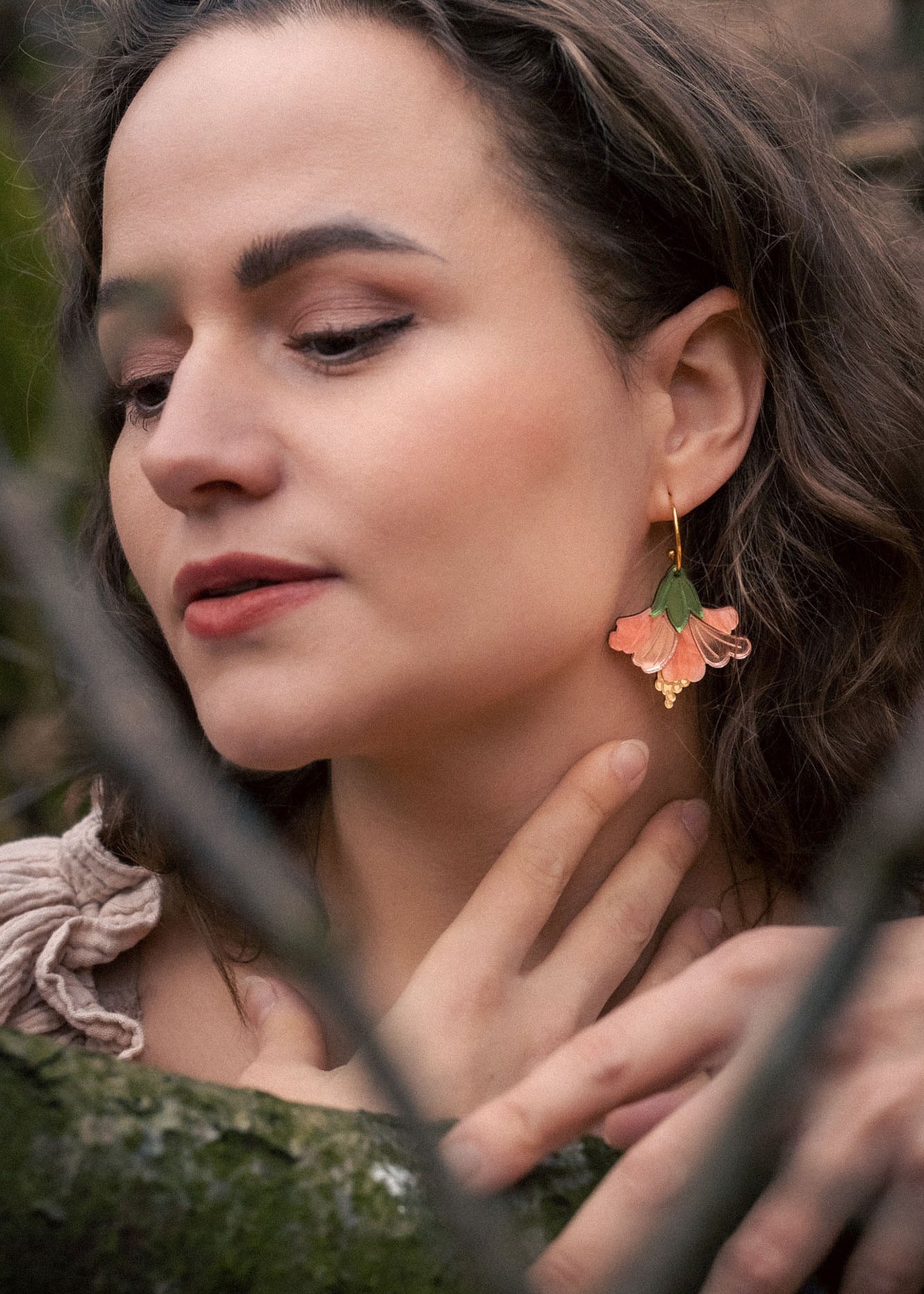 A woman with wavy brown hair and handcrafted Hibiscus Earrings gently touches her neck while standing in a forest. Her eyes are closed, branches cross before her, and she wears a beige top with ruffled details.