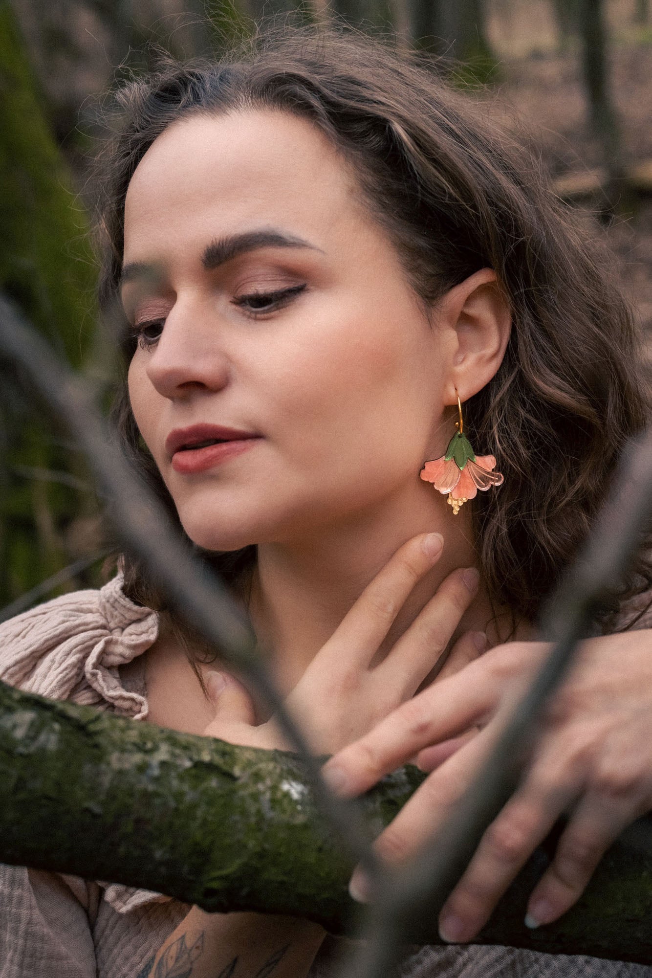 A woman with wavy brown hair and handcrafted Hibiscus Earrings gently touches her neck while standing in a forest. Her eyes are closed, branches cross before her, and she wears a beige top with ruffled details.