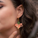 Close-up of a woman with wavy brown hair wearing handcrafted Hibiscus Earrings shaped like a pink and green flower, touching her neck gently with her fingers for a tropical statement look.
