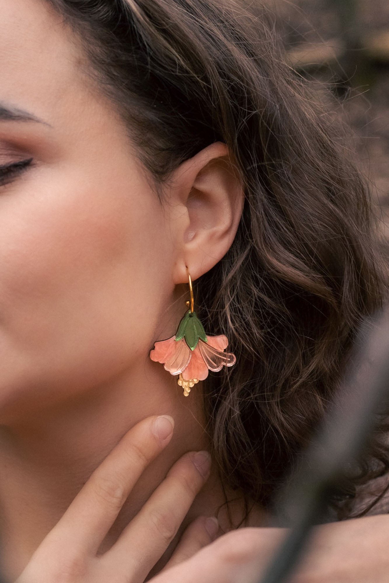 Close-up of a woman with wavy brown hair wearing handcrafted Hibiscus Earrings shaped like a pink and green flower, touching her neck gently with her fingers for a tropical statement look.