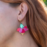 A close-up of a womans ear wearing handcrafted, petal-shaped Hibiscus Earrings in vibrant pink, teal, and gold tones. Her wavy, reddish-brown hair is partially visible, with tropical greenery softly blurred in the background.