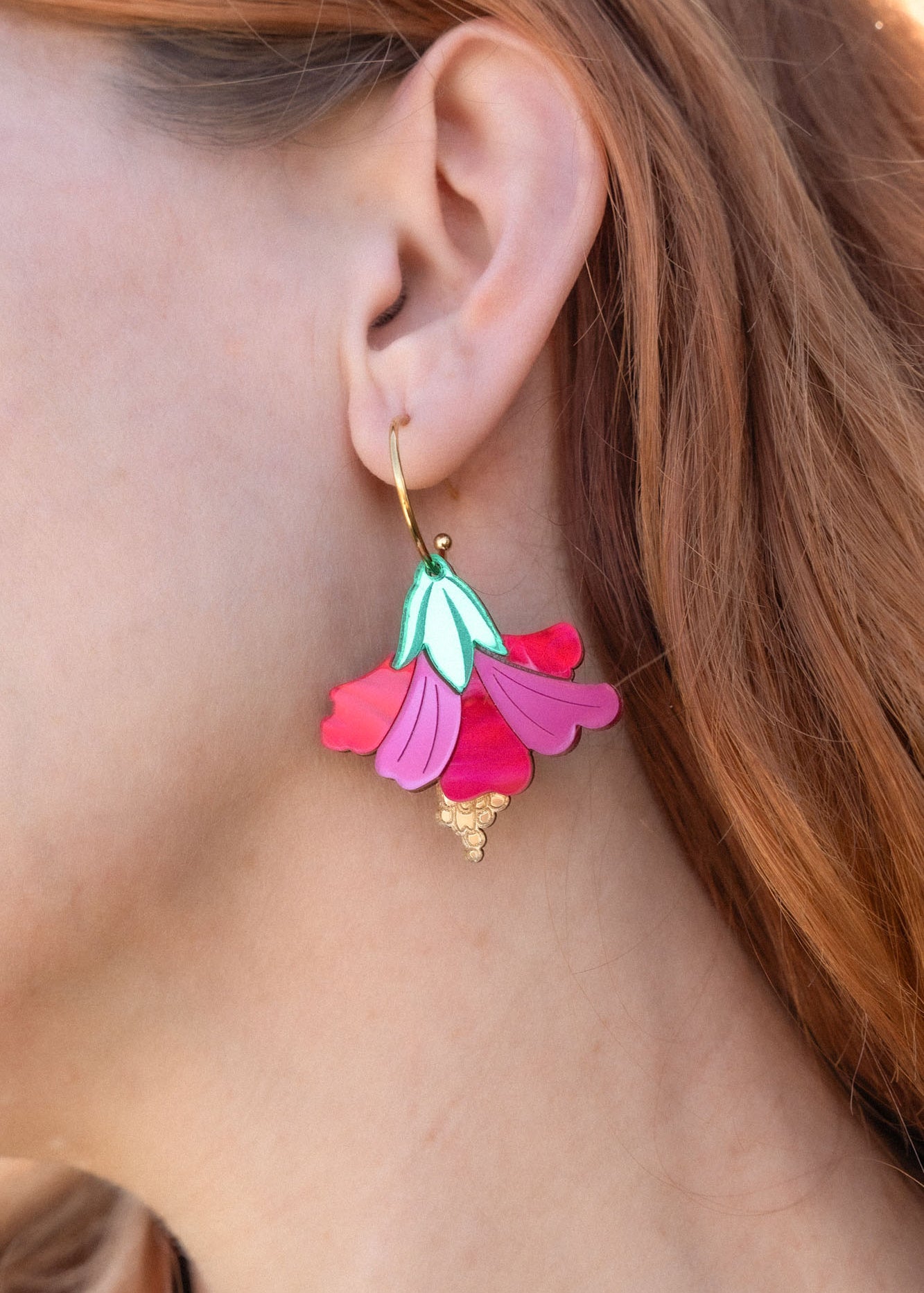 A close-up of a womans ear wearing handcrafted, petal-shaped Hibiscus Earrings in vibrant pink, teal, and gold tones. Her wavy, reddish-brown hair is partially visible, with tropical greenery softly blurred in the background.
