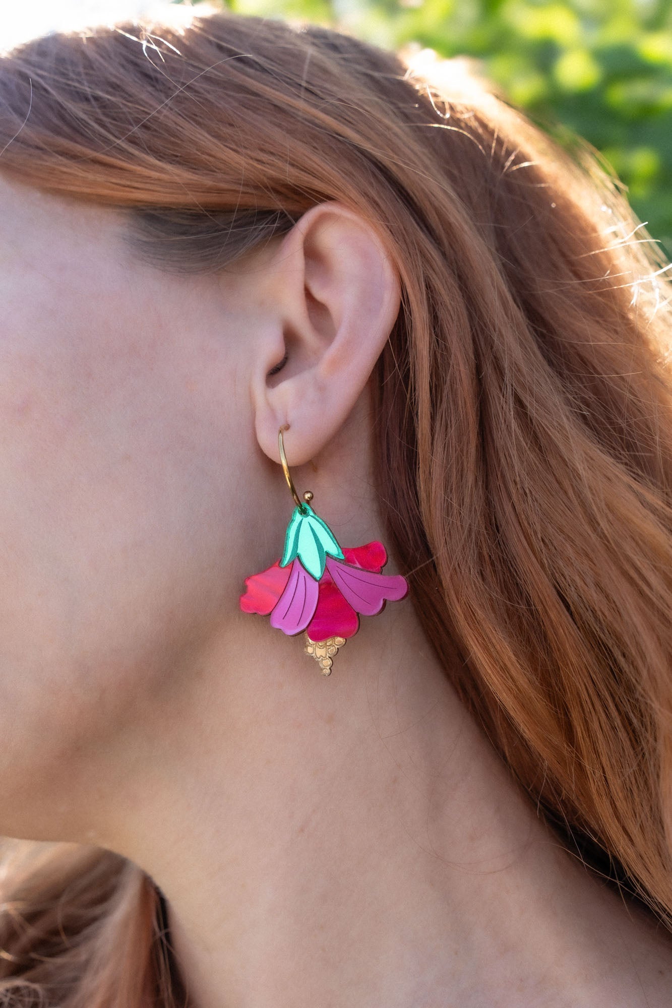 A close-up of a womans ear wearing handcrafted, petal-shaped Hibiscus Earrings in vibrant pink, teal, and gold tones. Her wavy, reddish-brown hair is partially visible, with tropical greenery softly blurred in the background.