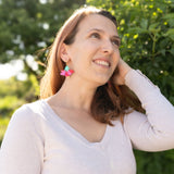 A woman with light brown hair smiles and looks upward, standing outdoors in sunlight. She wears a light long-sleeve top and handcrafted Hibiscus Earrings, with greenery and trees in the background.