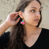 A woman with long brown hair wearing a black top and handcrafted, colorful Hibiscus Earrings looks to the side, touching her ear. The background is a light, textured wall.