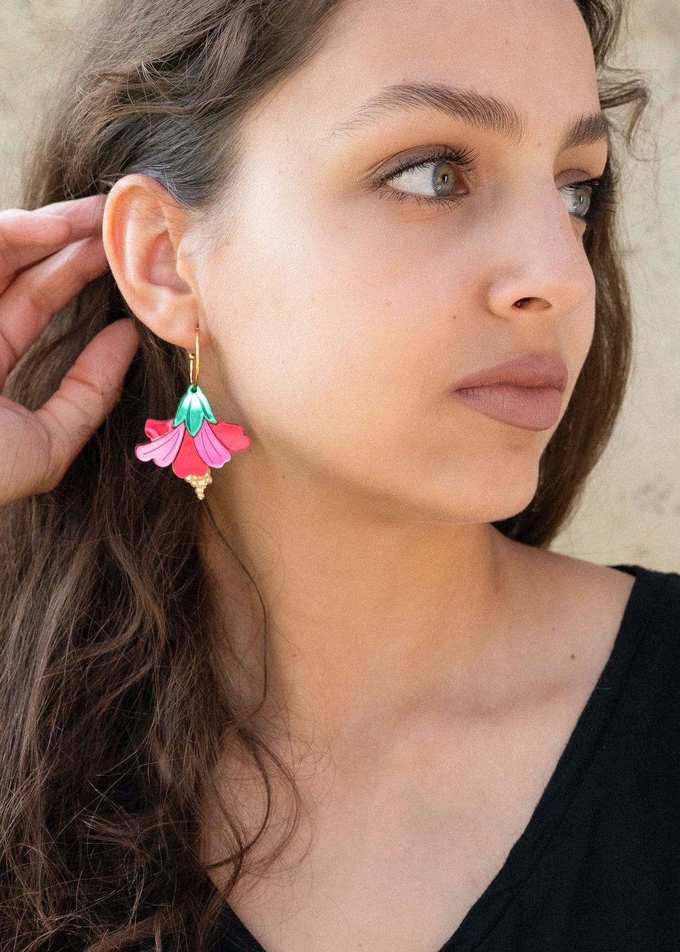 A woman with long brown hair wearing a black top and handcrafted, colorful Hibiscus Earrings looks to the side, touching her ear. The background is a light, textured wall.