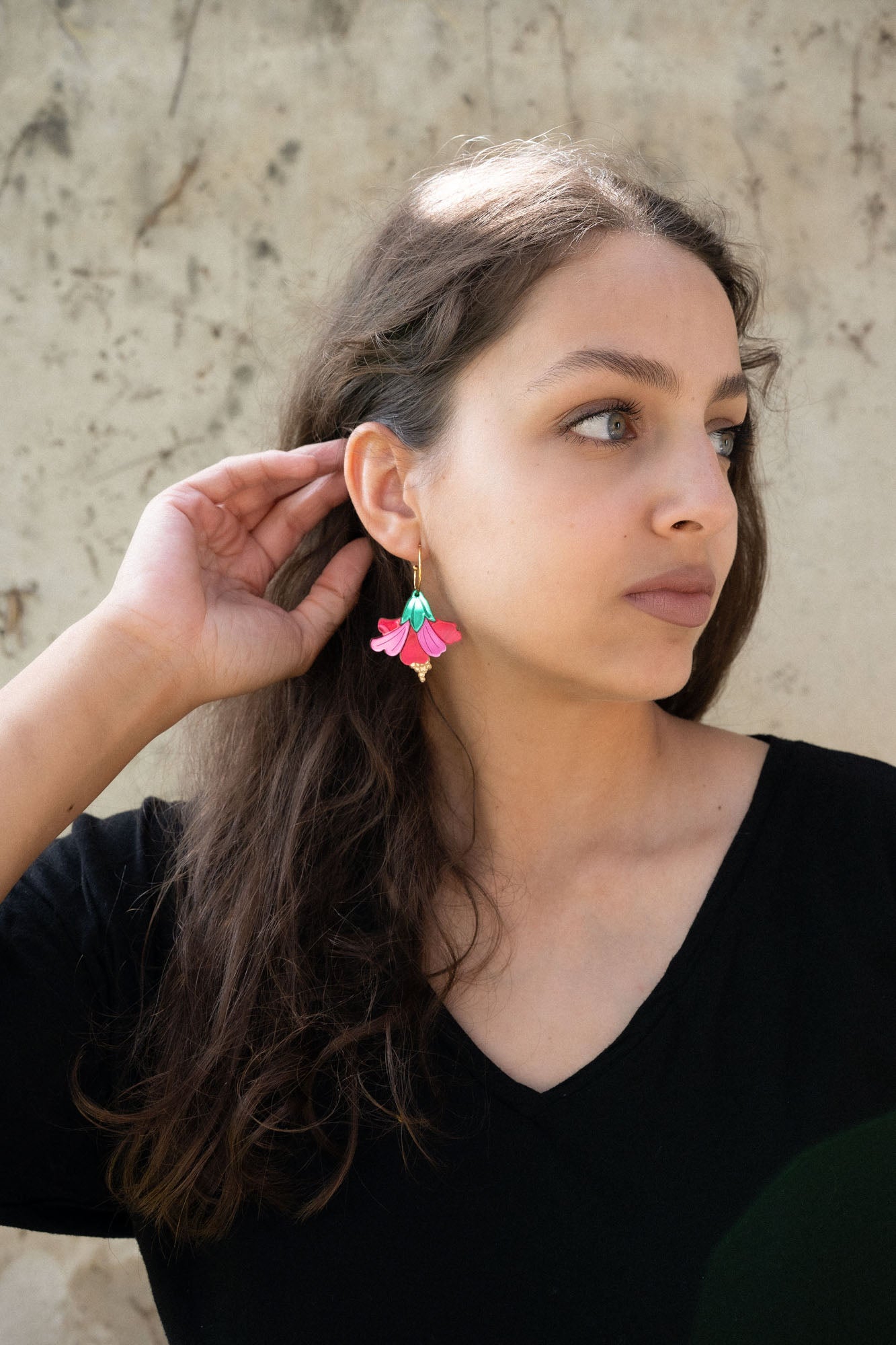 A woman with long brown hair wearing a black top and handcrafted, colorful Hibiscus Earrings looks to the side, touching her ear. The background is a light, textured wall.