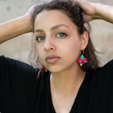 A young woman with light eyes and wavy brown hair poses with her hands behind her head, wearing a black top and handcrafted, colorful Hibiscus Earrings, looking directly at the camera against a neutral background.
