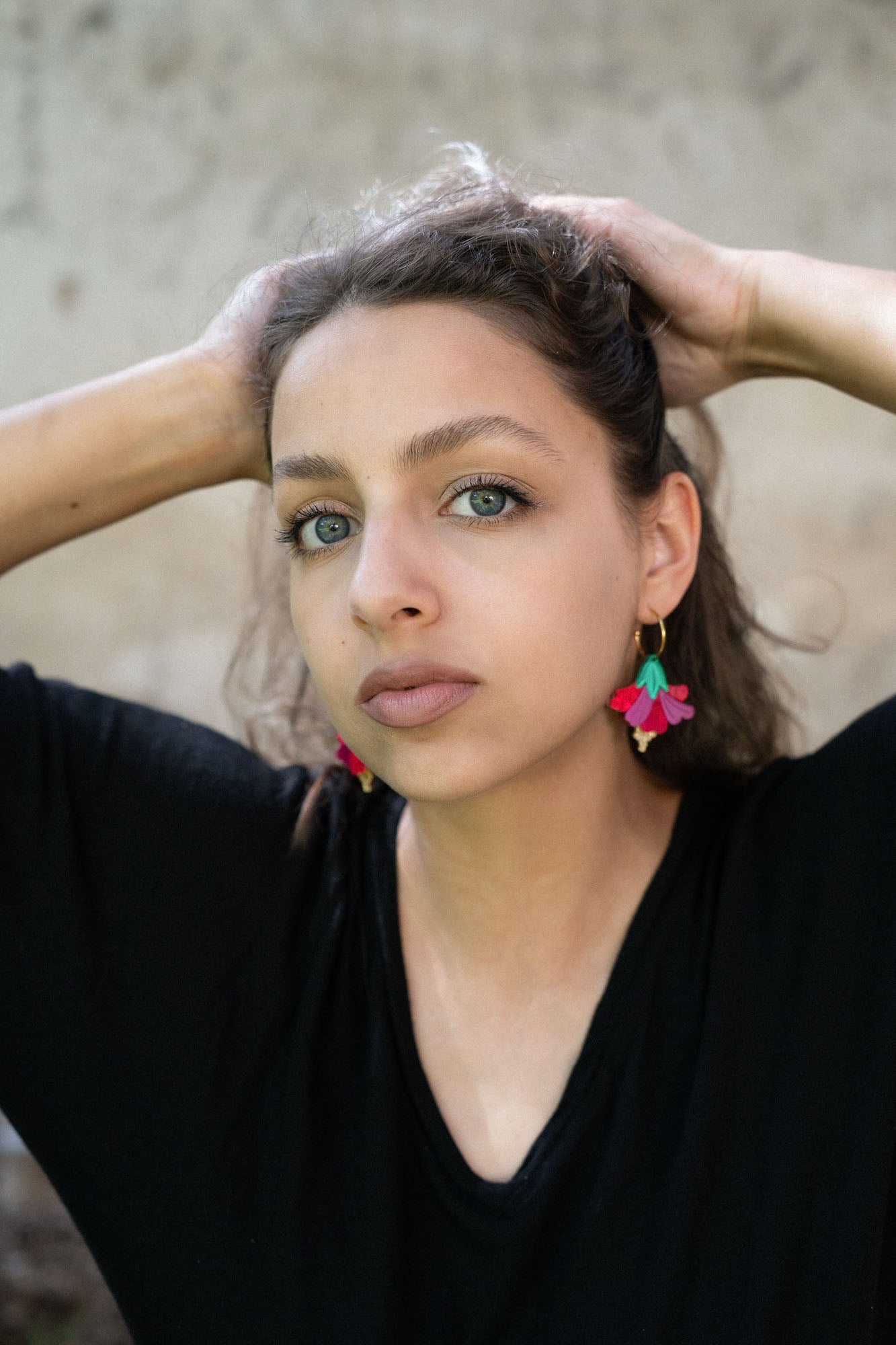A young woman with light eyes and wavy brown hair poses with her hands behind her head, wearing a black top and handcrafted, colorful Hibiscus Earrings, looking directly at the camera against a neutral background.