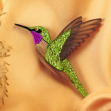 A handcrafted hummingbird brooch with vibrant green and purple glittery hues and brown wings rests on soft, peach-colored fabric.