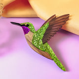 A handcrafted Hummingbird Brooch with vibrant colors—glittery green wings, a purple head, and brown accents—rests on a pastel pink surface with a yellow fabric backdrop.