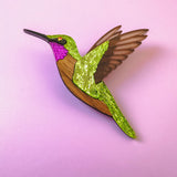 A handcrafted Hummingbird Brooch with vibrant colors—green, pink, brown, and gold glitter accents—shines beautifully against a light pink background.
