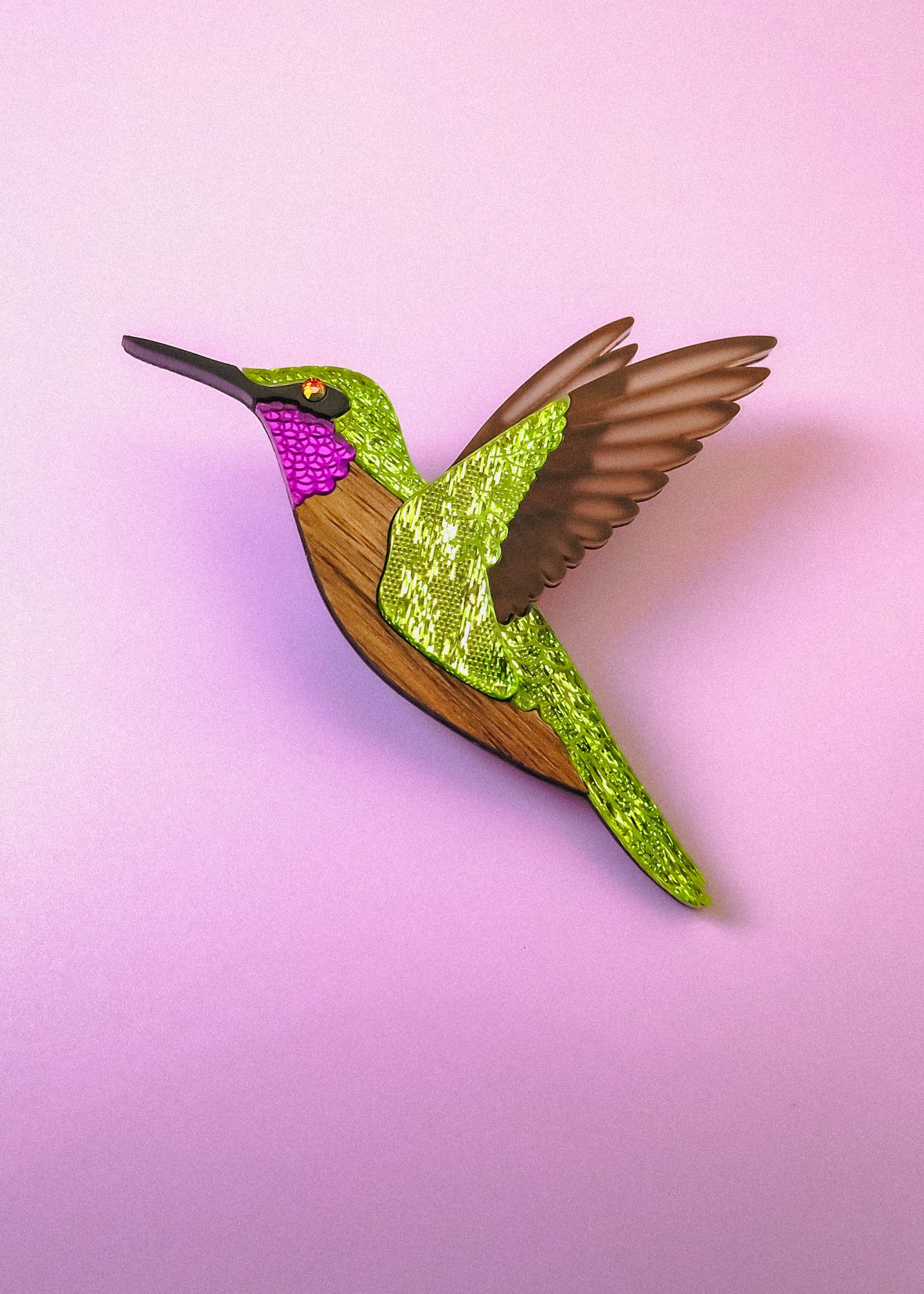 A handcrafted Hummingbird Brooch with vibrant colors—green, pink, brown, and gold glitter accents—shines beautifully against a light pink background.