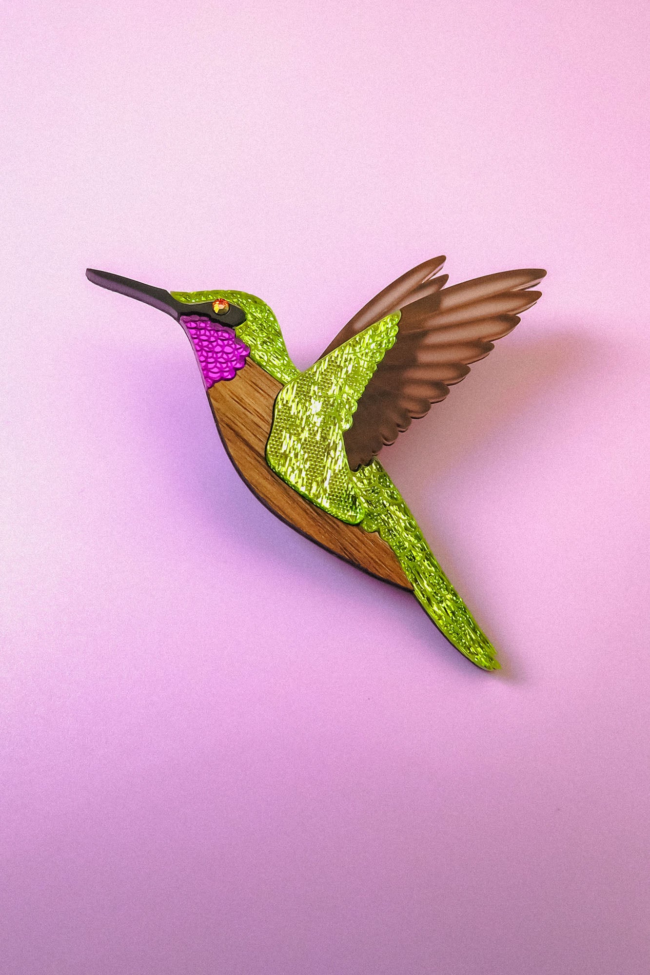 A handcrafted Hummingbird Brooch with vibrant colors—green, pink, brown, and gold glitter accents—shines beautifully against a light pink background.