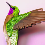A close-up of a handcrafted hummingbird brooch in vibrant colors—metallic green and purple with brown wings—set against a soft pink background.