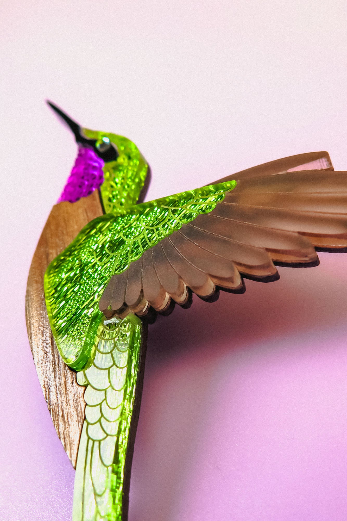 A close-up of a handcrafted hummingbird brooch in vibrant colors—metallic green and purple with brown wings—set against a soft pink background.