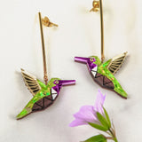 A pair of handcrafted gold and colorful geometric Hummingbird Earrings lie on a white fabric background, with a small pink flower in the foreground.