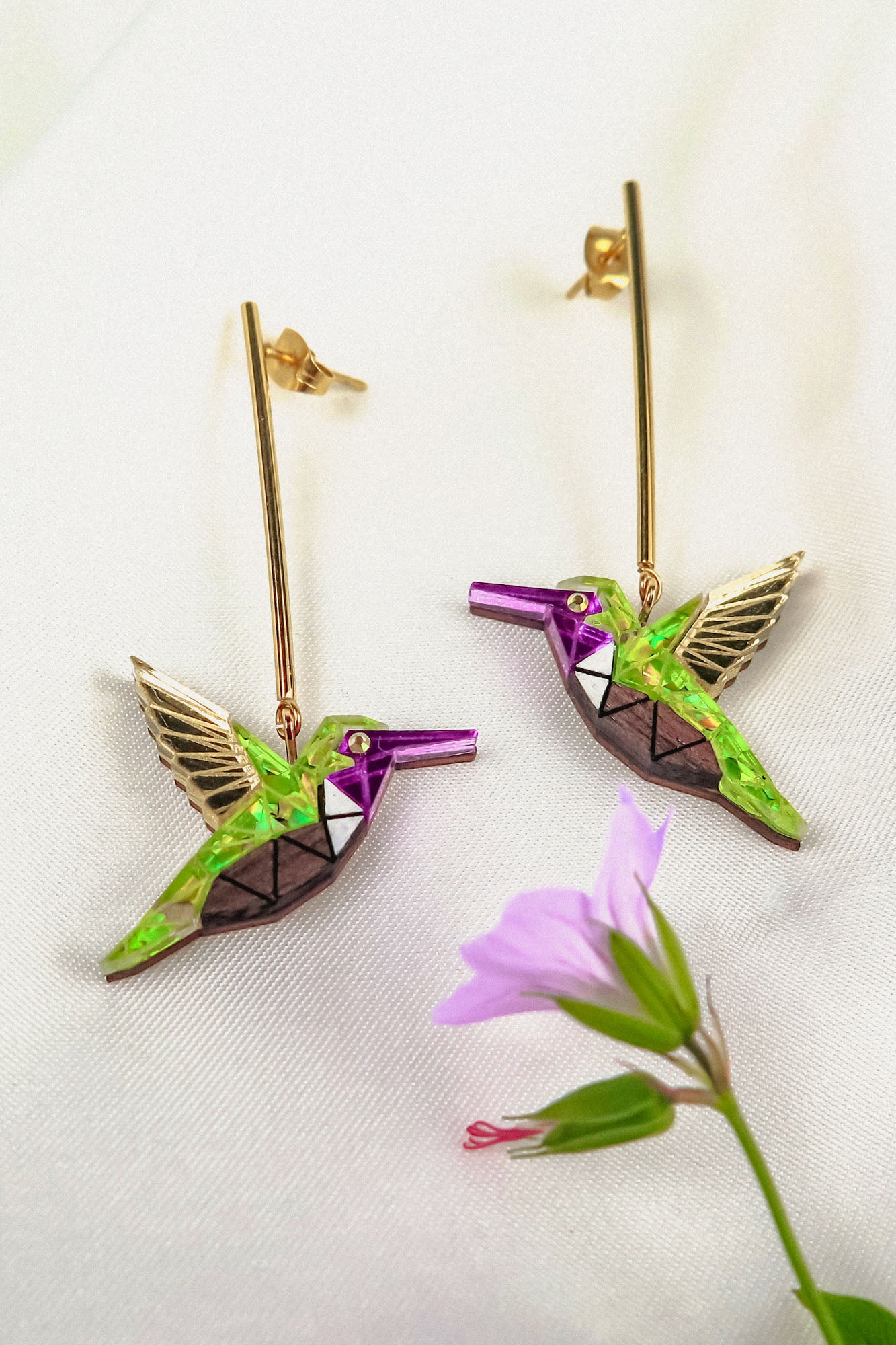 A pair of handcrafted gold and colorful geometric Hummingbird Earrings lie on a white fabric background, with a small pink flower in the foreground.